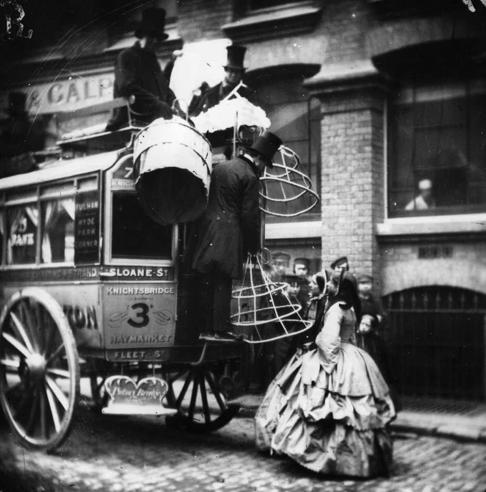 Victorian-era street scene with people in formal attire boarding a horse-drawn carriage. Women in large dresses and men in top hats interact, with a backdrop of brick buildings. Passersby observe the scene.