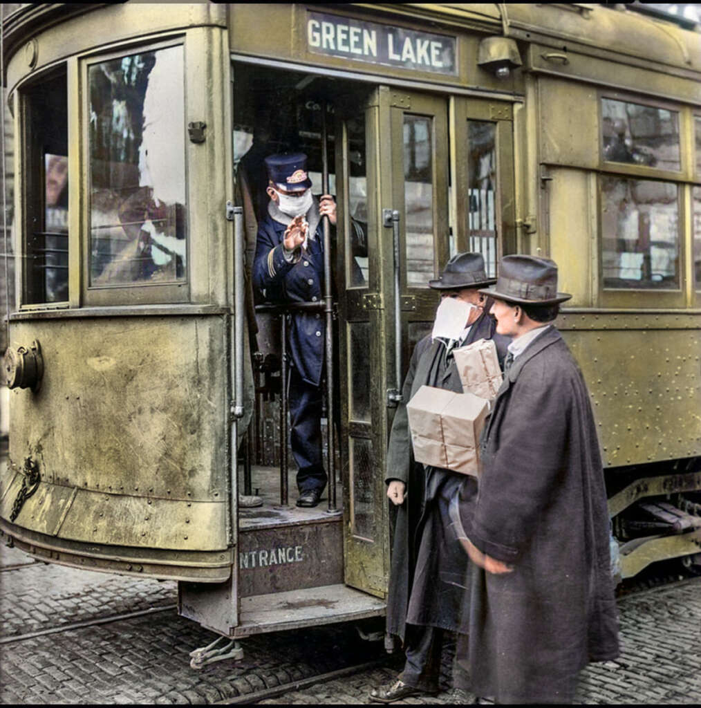 A historic trolley marked "Green Lake" with a conductor signaling, wearing a uniform and mask. Two men in early 20th-century attire and masks stand outside; one holds a paper bag. Cobblestone street visible beneath.