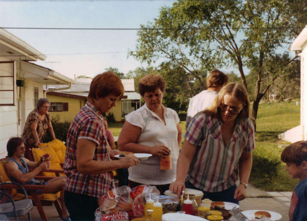 A group of people gather outdoors for a casual meal. Two women in plaid shirts prepare plates at a table with food items, while others sit and stand nearby. Trees and houses are visible in the background.