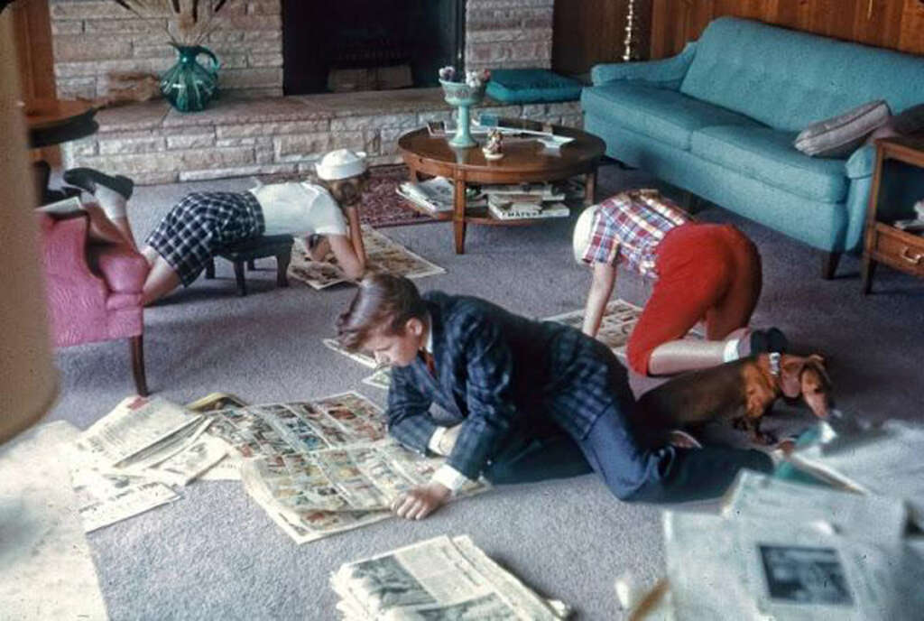 Three children are lying on a carpeted floor, reading comic sections of newspapers. A dog is resting beside them. The room features a blue sofa, coffee table with items, and a brick fireplace with vase.