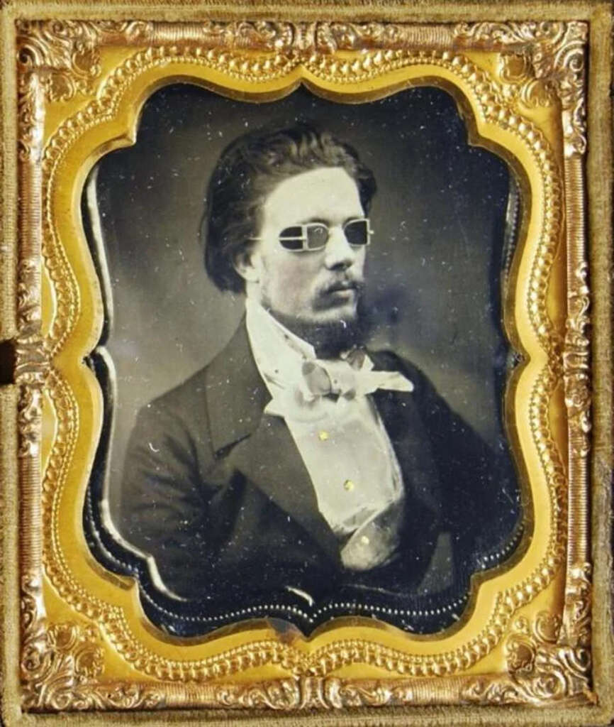 A vintage photograph of a man with sunglasses, wearing a formal suit and bow tie. The image is framed in an ornate gold border, typical of 19th-century portrait styles.