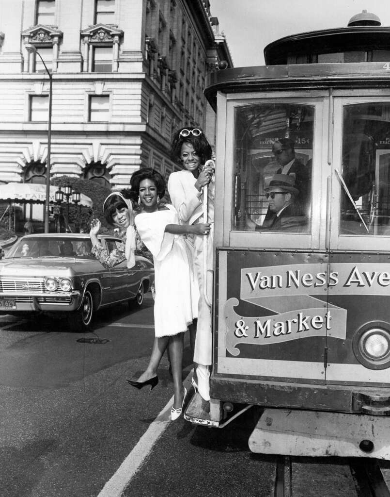 Three women in 1960s attire joyfully ride on the side of a vintage San Francisco cable car labeled "Van Ness Ave & Market." One woman in a car waves in the background. The scene captures a lively street with vintage cars and historic buildings.