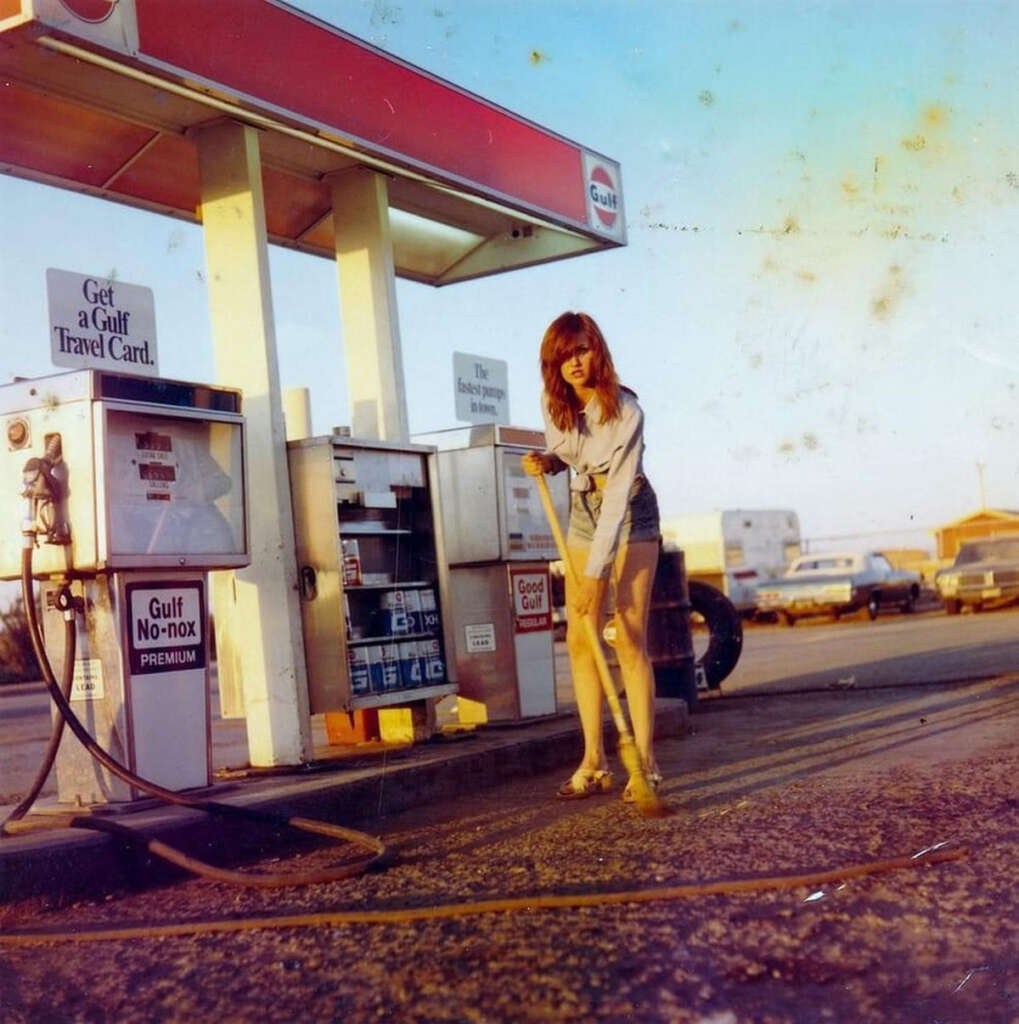 A woman wearing a light-colored top and shorts is using a squeegee on the ground at a vintage Gulf gas station. There's a gas pump on her left and several cars parked in the background under a clear sky.