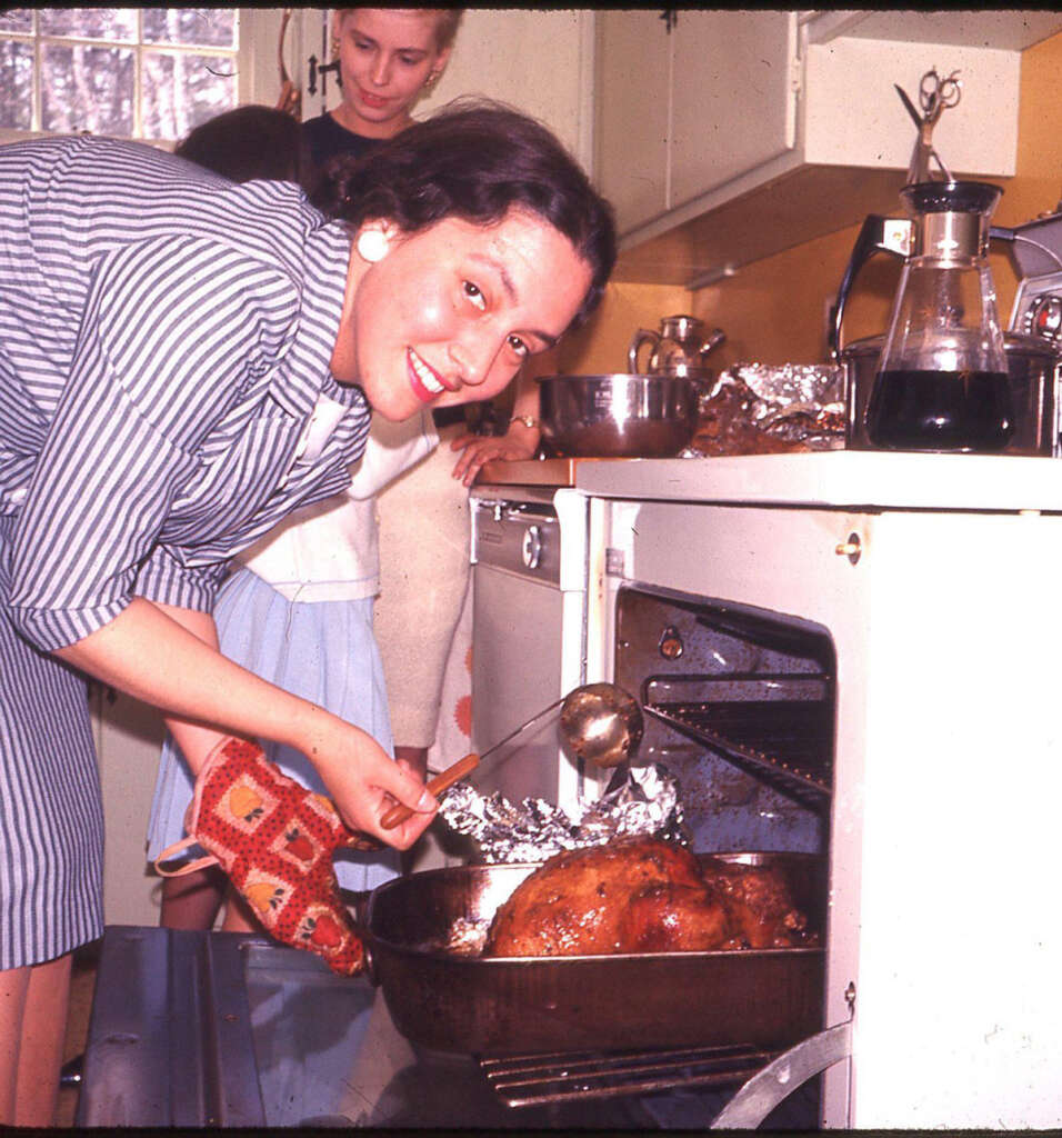 A person smiling at the camera while basting a roasted turkey in an oven. Another person stands behind, watching, in a kitchen setting with yellow walls and a coffee pot on the counter. The main person is wearing a striped outfit and holding an oven mitt.