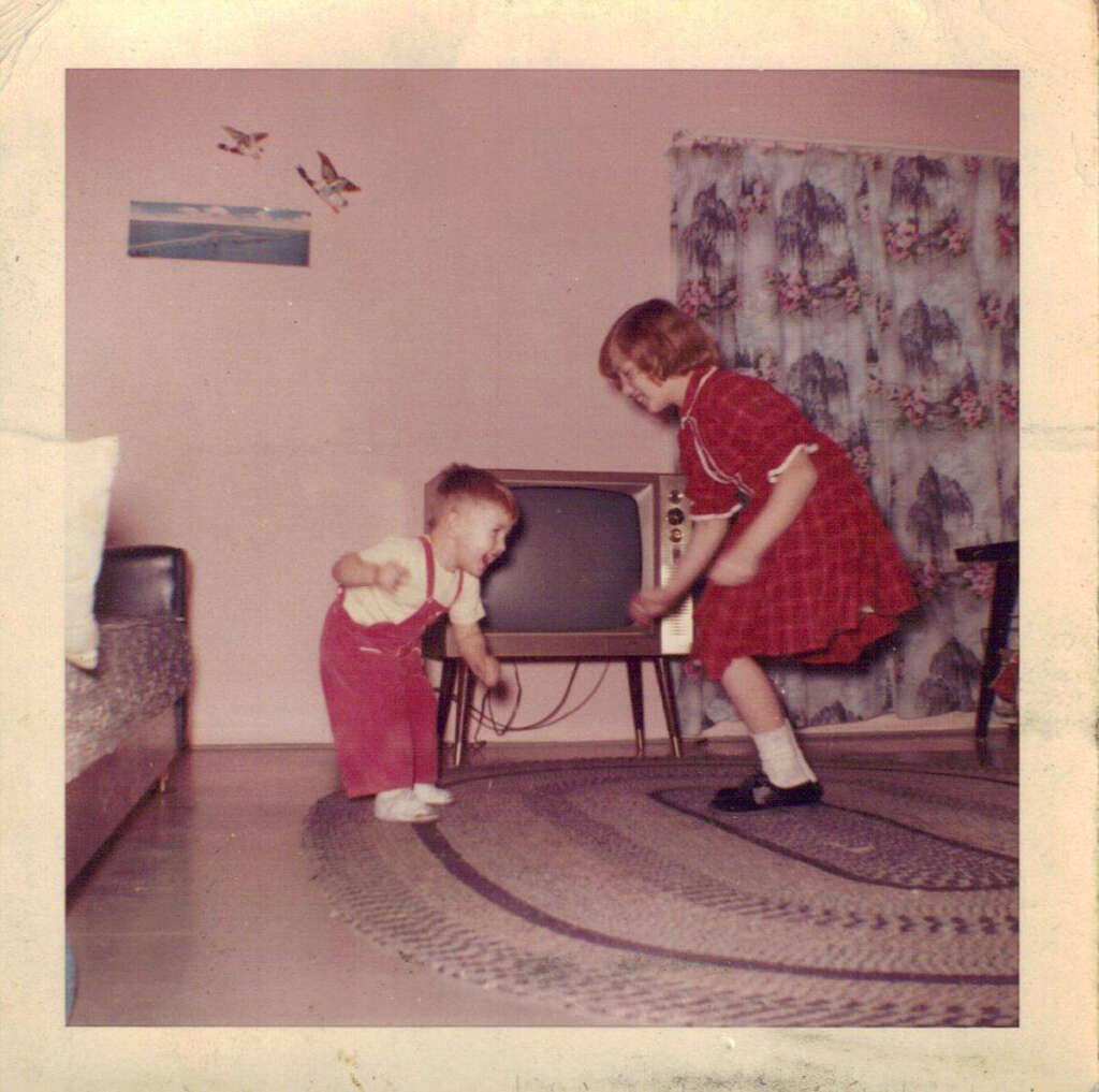 Two children play in a living room. A younger child in red overalls holds a toy, while an older child in a red dress interacts with them. A vintage television sits in the background, and floral curtains adorn the window. The room has a large rug.