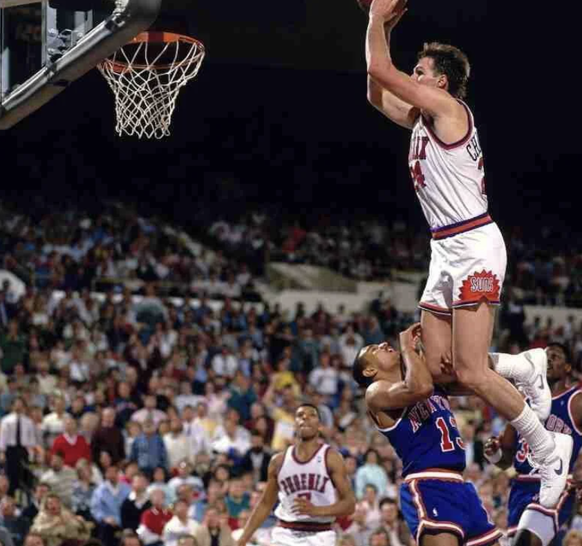 A basketball player in a white Phoenix Suns uniform is jumping high near the basket, preparing to score. An opposing player in a blue jersey is attempting to block. The background shows a large crowd watching the game.