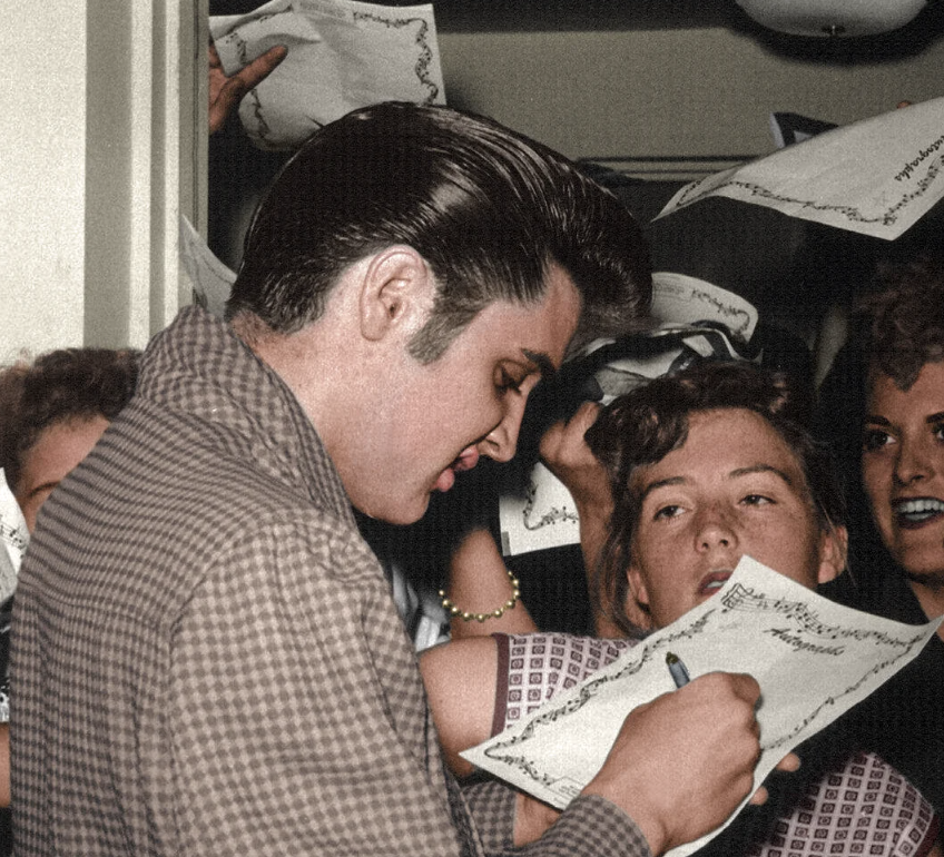 A man with slicked-back hair in a checkered jacket signs autographs for a group of excited fans, who are holding out papers. The scene appears lively with smiles and eager expressions.