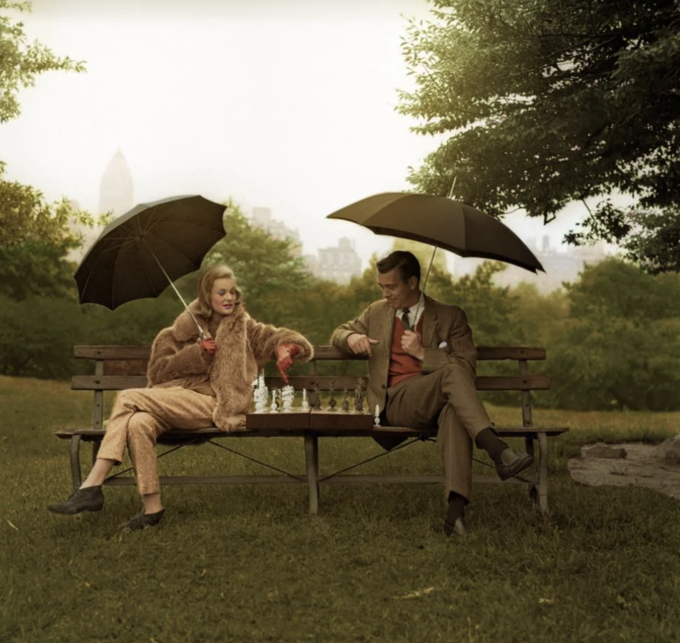 Two people sit on a park bench playing chess. Both hold umbrellas. The person on the left, wearing a fur coat, holds a black umbrella. The person on the right, in a suit, holds a larger black umbrella. The city skyline is visible in the foggy background.