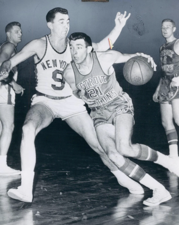 A vintage basketball game scene with a player from Detroit Pistons dribbling the ball while a New York Knicks player tries to block him. Other players are visible in the background. The photo is in black and white.