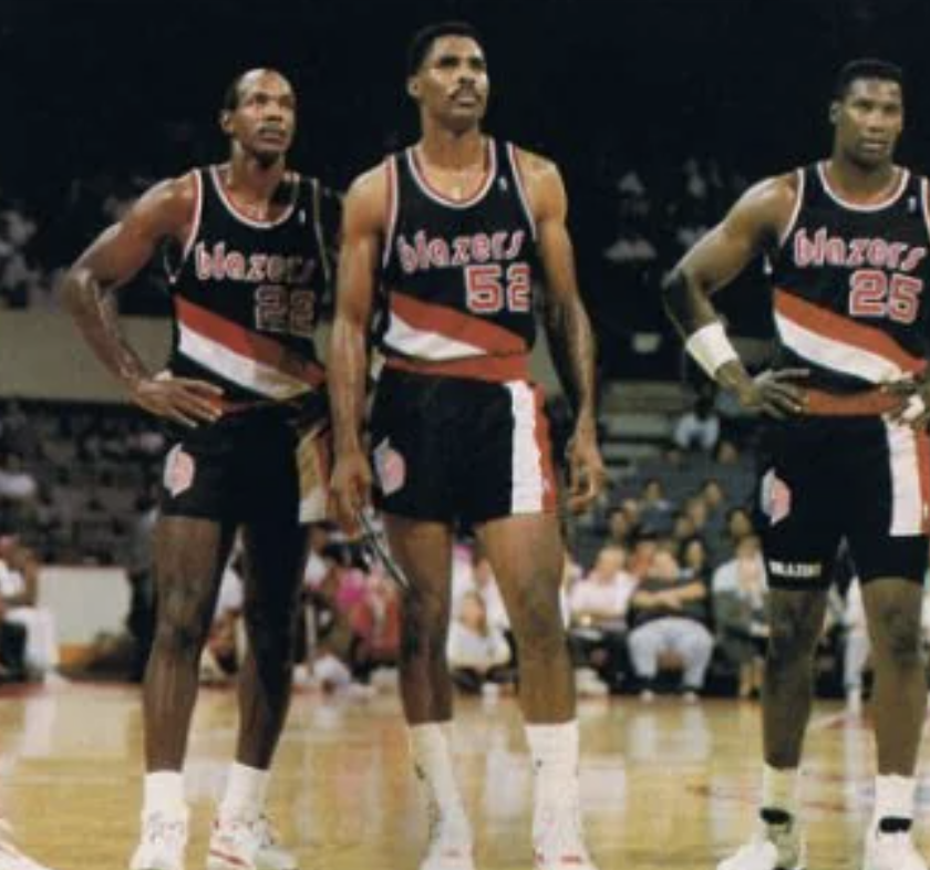 Three basketball players in black "Blazers" uniforms stand together on the court, with hands on hips. The number 52 is in the center, 22 on the left, and 25 on the right. Spectators are visible in the background.