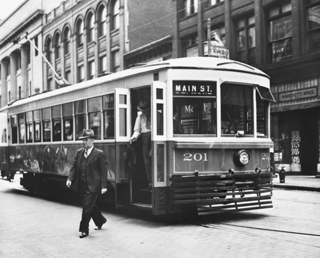 A vintage streetcar marked "Main St." is stopped on a city street. A man in a suit and hat walks in front of it, and another person is stepping off. Historic brick buildings line the background. The scene appears to be from the early 20th century.