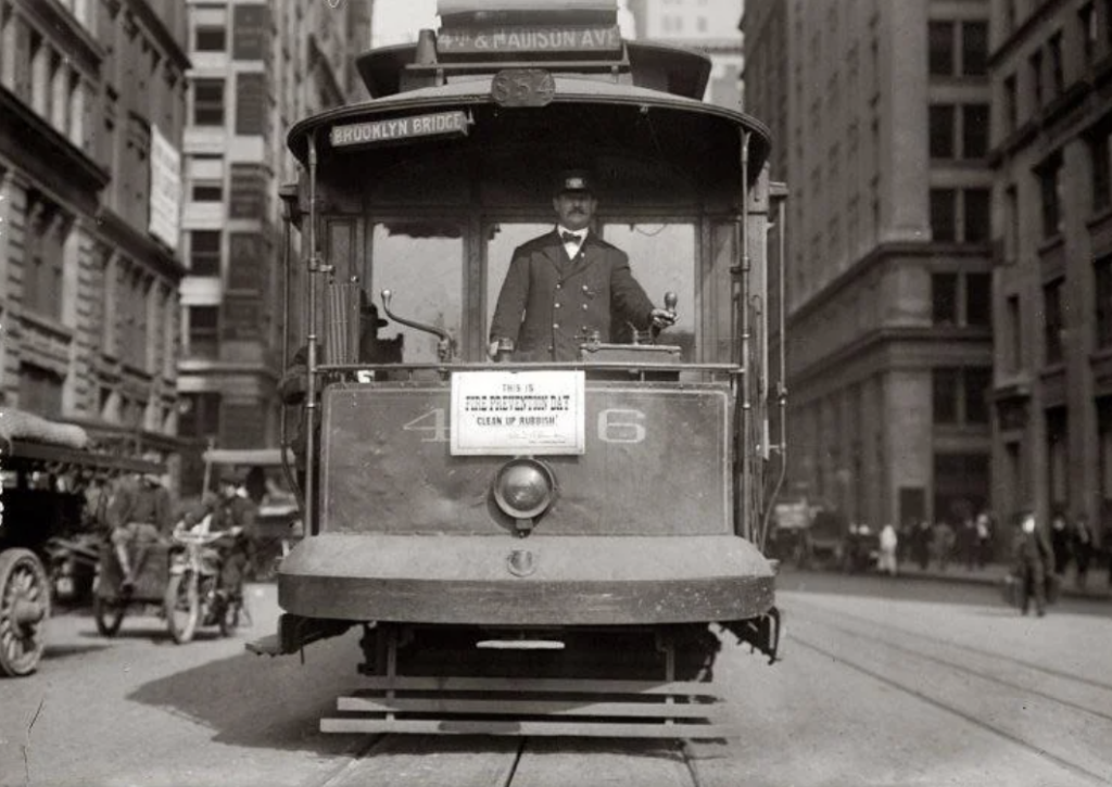 A black-and-white photo shows a vintage streetcar labeled "Brooklyn Bridge" on a city street. A conductor stands at the front, and horse-drawn carriages and pedestrians can be seen in the background among tall buildings.