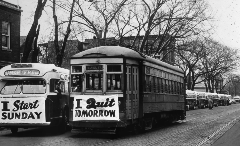 A vintage street scene shows a streetcar with a sign reading "I Quit Tomorrow" and a bus with a sign reading "I Start Sunday." Bare trees line the street, and multiple buses are visible in the background.