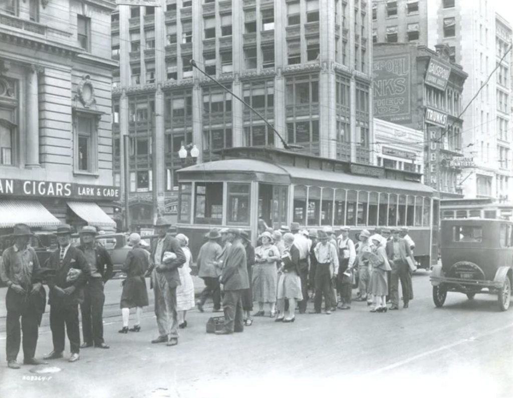 A black and white photo of a street scene with a streetcar in the center. Many people are gathered near the streetcar, and early 20th-century buildings surround them. There are vintage cars and large signs on the buildings.