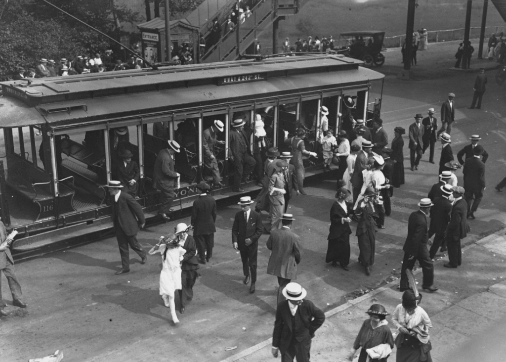 A black and white photo depicts a bustling street scene with people in early 20th-century attire boarding and exiting a streetcar. Men and women wear hats and suits, while a few hold umbrellas. The background shows a staircase and a sparse crowd.