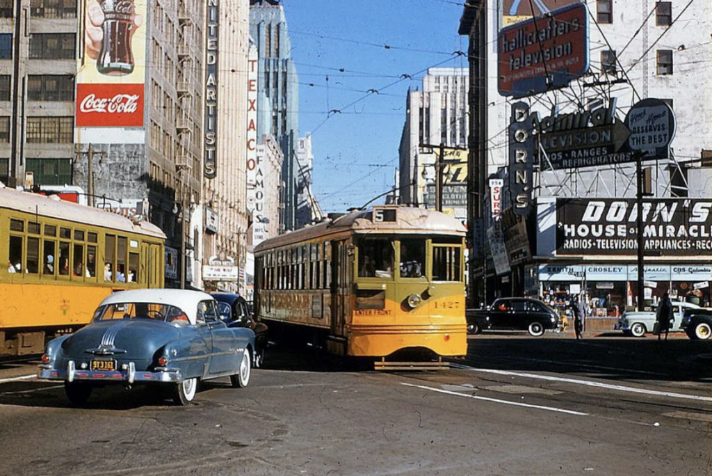 A vintage city street scene with yellow streetcars and a blue classic car. Tall buildings feature signs for Coca-Cola and various businesses. Pedestrians are visible on the sidewalks under clear skies.