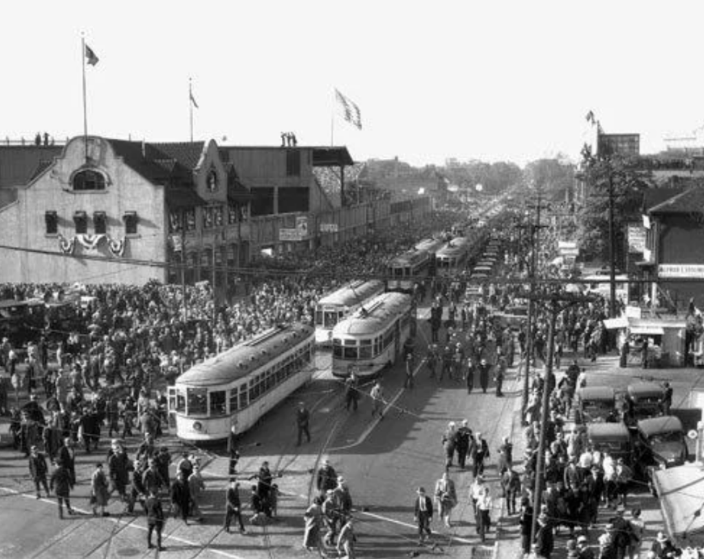 Black and white photo of a bustling street scene with numerous people around streetcars. A large crowd fills the street, flanked by buildings adorned with flags and banners. Vintage cars are parked along the curb, and there is a stadium in the background.