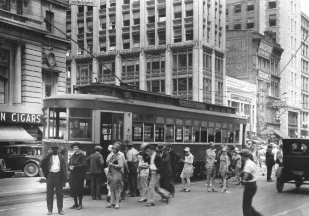 Black and white photo of people boarding and disembarking a streetcar in a bustling city scene from the past. The street is busy with pedestrians and cars, and there are tall buildings and various signage in the background.