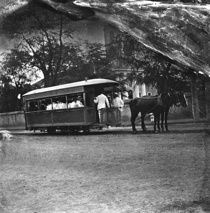 A vintage image depicts a horse-drawn tramcar on a street, with people standing inside. Trees and a building are visible in the background. The photo has an aged and textured appearance.