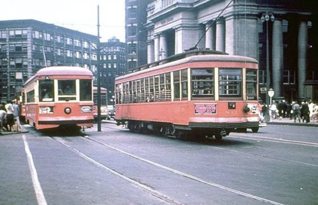 Historic streetcars painted in red and beige travel on tracks in a bustling urban area with tall buildings and a crowd of pedestrians. One streetcar is approaching while the other is turning.