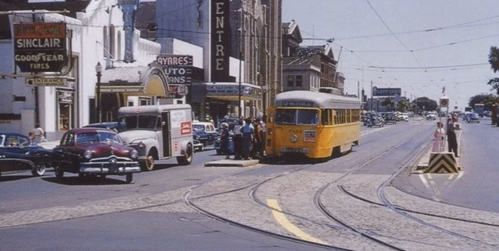 Vintage street scene with a yellow streetcar on tracks, surrounded by 1950s-era cars. A group of people wait near a gas station displaying "Sinclair" and "Goodyear Tires" signs. Buildings line the sunny street, with overhead tram wires visible.