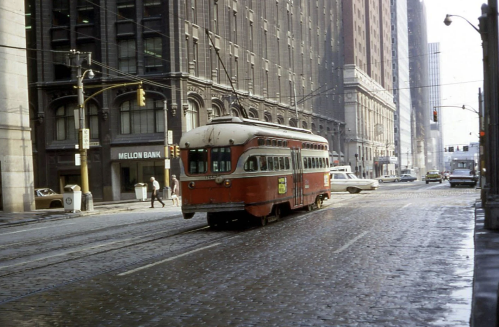 A vintage red streetcar travels along a cobblestone street between tall buildings. To the left, a Mellon Bank branch is visible. Cars and pedestrians move along the wet street as the city looms in the background.