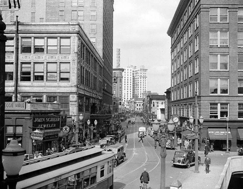 Black and white photo of a busy urban street scene from the mid-20th century. Vintage cars and a streetcar are visible. Pedestrians walk along sidewalks beside buildings with prominent signs for a jeweler and other shops.