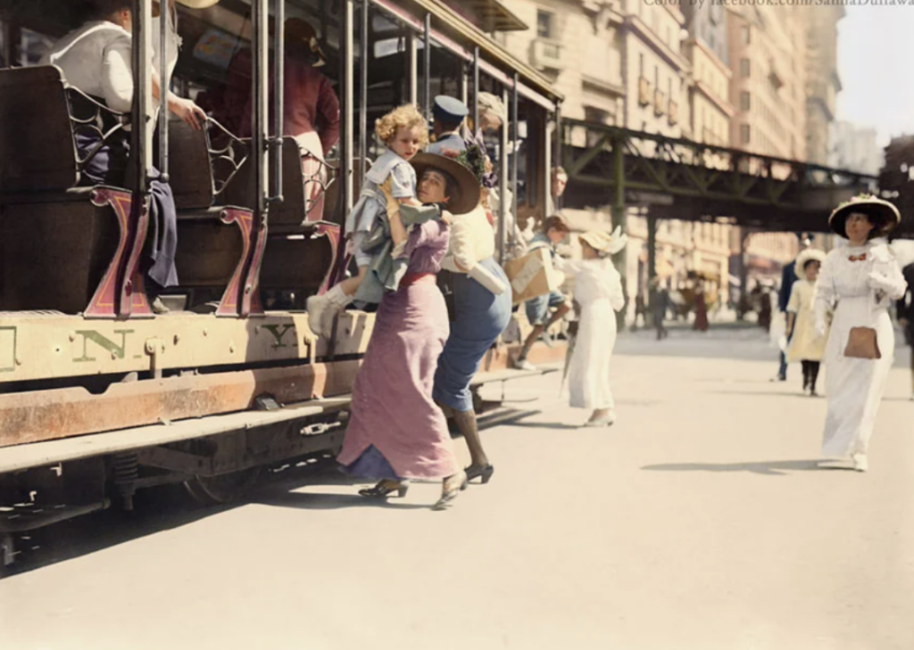 A vintage street scene with a woman helping a child onto a crowded trolley car. The woman wears a long dress and hat, while the bustling city street shows people walking, and a blurred elevated train track in the background.