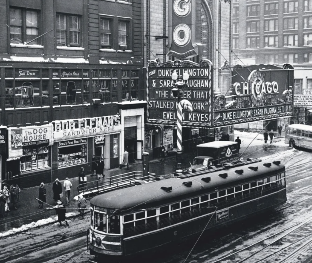 A vintage street scene features a tram passing through a busy urban area. People walk on the sidewalks near shops like a sandwich shop and a dog house eatery. Marquees advertise entertainers and movies, with a backdrop of tall buildings and light snow.