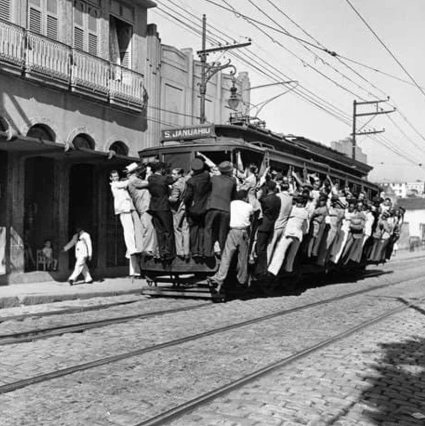 A vintage black and white photo shows a crowded streetcar with numerous passengers hanging onto the sides. The tram moves along cobblestone tracks in a street lined with buildings, capturing a bustling, busy scene.