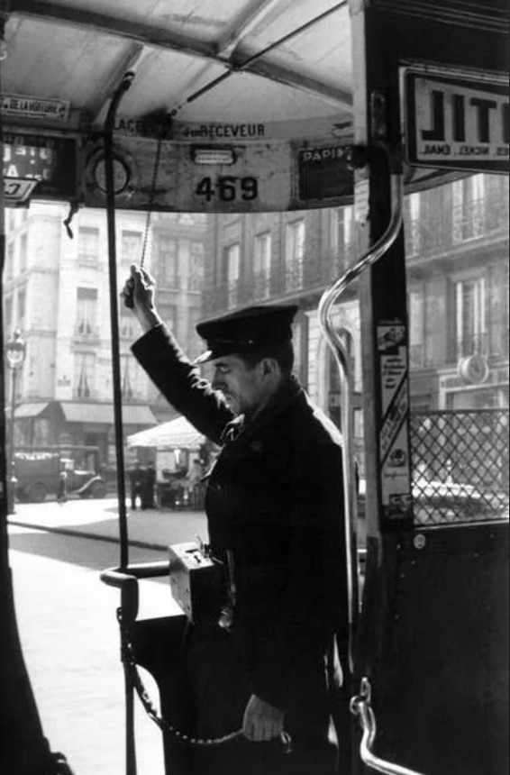 A vintage photo of a uniformed ticket collector standing in a tram, holding a cord. He wears a cap and a ticket bag. The tram interior shows signage and advertisements. The background reveals an urban street scene with buildings.