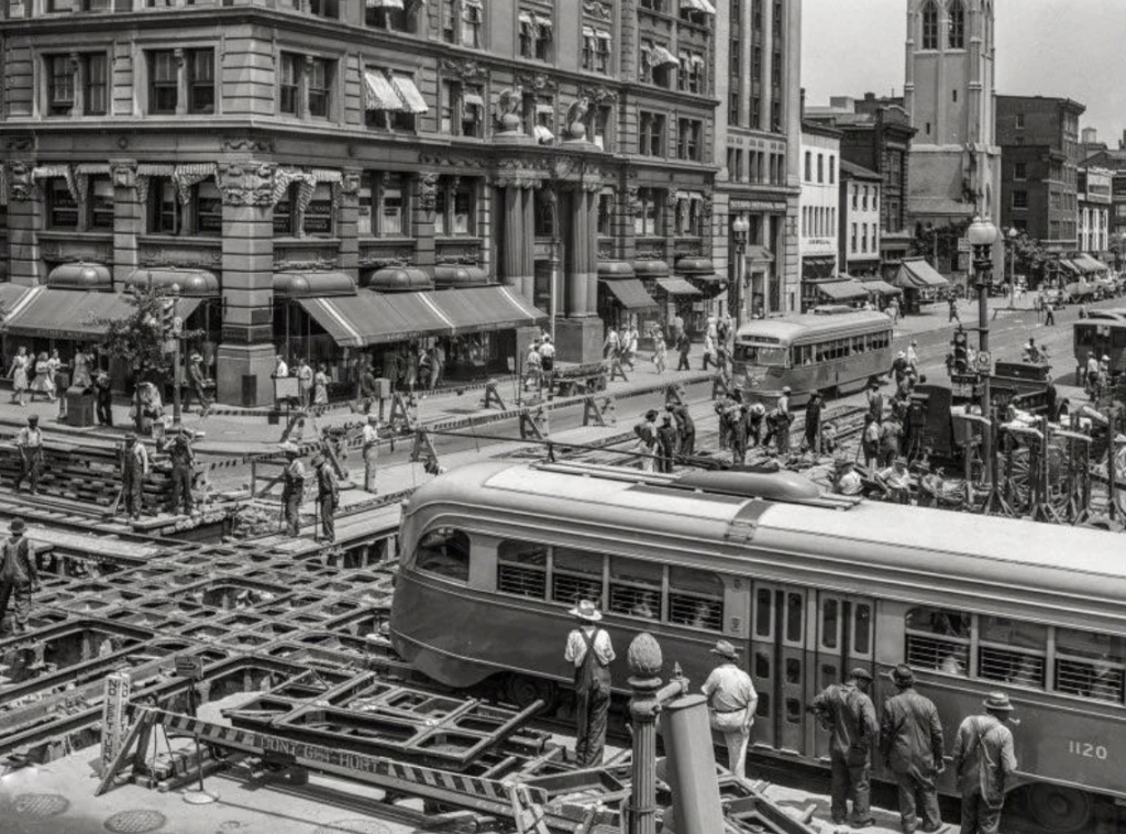 Black and white photo of a bustling city scene from the mid-20th century. Workers are laying streetcar tracks on a busy intersection surrounded by historic buildings. People walk on sidewalks and cars are visible in the background.