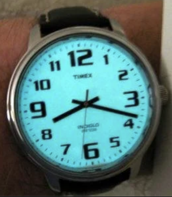 A Timex wristwatch with a glowing Indiglo dial shows the time as 10:09. The watch has a silver case, black numerals, and a black leather strap.