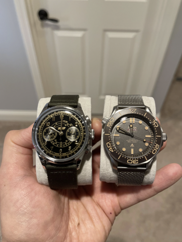 A hand holds two watches on stands: the left watch has a black face with multiple dials and a dark strap; the right watch features a gray face with a stainless steel mesh band, styled like a diving watch. A white door is in the background.