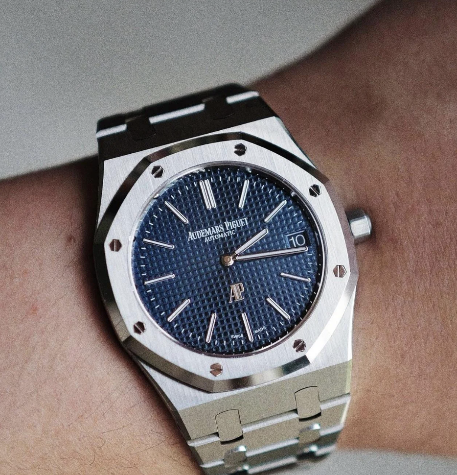 A silver Audemars Piguet wristwatch with a textured blue face is shown on a person's wrist. The watch features a metal link band, a date display, and small silver hour markers and hands.