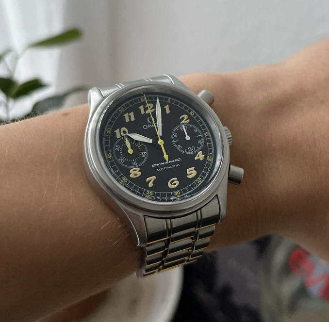 A person wearing a silver wristwatch with a black face and yellow numbers. The watch has three subdials and a metal band. The background is blurry, showing a hint of green plant leaves.