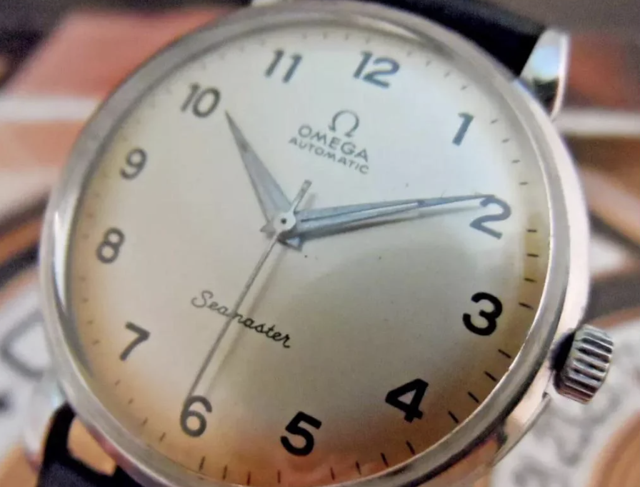 Close-up of a vintage Omega Seamaster watch with a silver face and black numerals. The watch features a simple design with three hands and the "Omega Automatic" logo at the top. The strap is black leather.