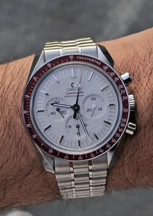 A close-up of a wrist with an Omega Speedmaster Professional watch. It features a white dial, red tachymetric scale bezel, and a stainless steel bracelet. The watch displays hours, minutes, seconds, and has three sub-dials.