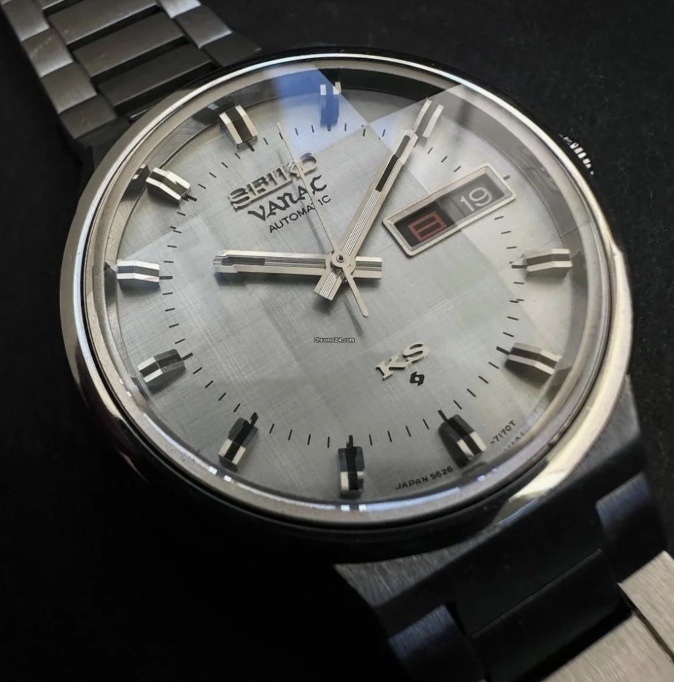 Close-up of a Seiko watch with a silver stainless steel band. The watch face features a date display set to August 19, with the day written in red. It has sleek metal hour markers and hands, with the words "Seiko," "Vanac," "Automatic," and "Japan 5626" visible.