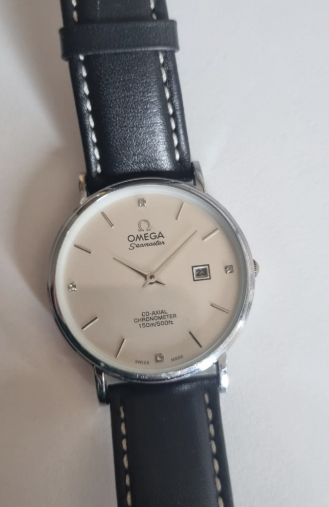 A silver Omega Seamaster watch with a white face and black leather strap. The watch features silver hour markers, a date window at the 3 o'clock position, and "Co-Axial Chronometer 150m/500ft" text on the dial.