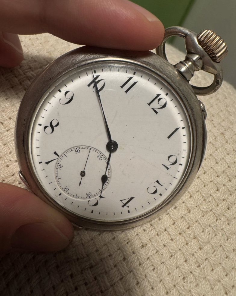 A silver pocket watch with black numbers and a smaller seconds dial is held by a hand. The watch features ornate hands, and the time reads approximately 10:09. The watch rests on a textured, cream-colored surface.