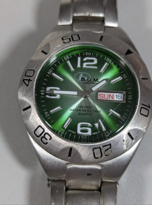 Close-up of a wristwatch with a metallic band, featuring a green dial. The watch shows the time as 1:50. It has bold white hour markers and hands. The date display reads "SUN 15." The brand name is visible on the dial.