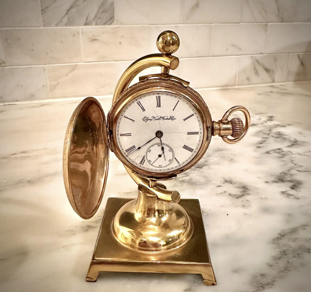 A vintage pocket watch mounted on an ornate, gold-toned stand. The watch face displays Roman numerals and a small second dial. Its cover is open. The stand rests on a marble surface with a tiled background wall.