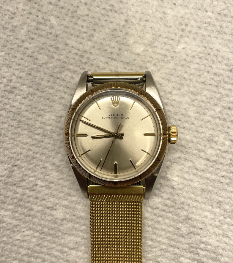 A Rolex Oyster Perpetual watch with a gold mesh strap is placed on a textured gray surface. The watch has a silver face with minimalist hour markers and a crown logo at the top.