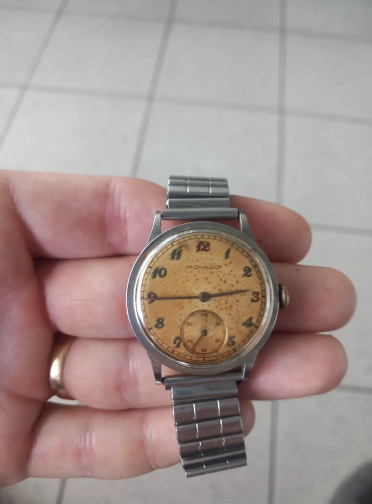 A hand holding a vintage Movado watch with a stainless steel band. The watch has a speckled, beige dial with black numerals and a small seconds subdial at the 6 o'clock position. The watch face shows signs of age.