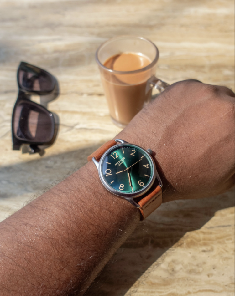 A person wearing a watch with a green face and tan strap rests their hand on a table. Nearby are a pair of sunglasses and a transparent mug filled with a light brown beverage. The table has a wood grain texture.