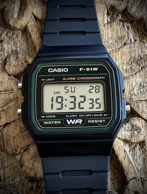 A close-up of a Casio F-91W digital wristwatch with a black strap placed on a textured wooden surface. The watch displays the time as 19:32:35 on Sunday, the 28th. It has a green and black face with multiple function labels.