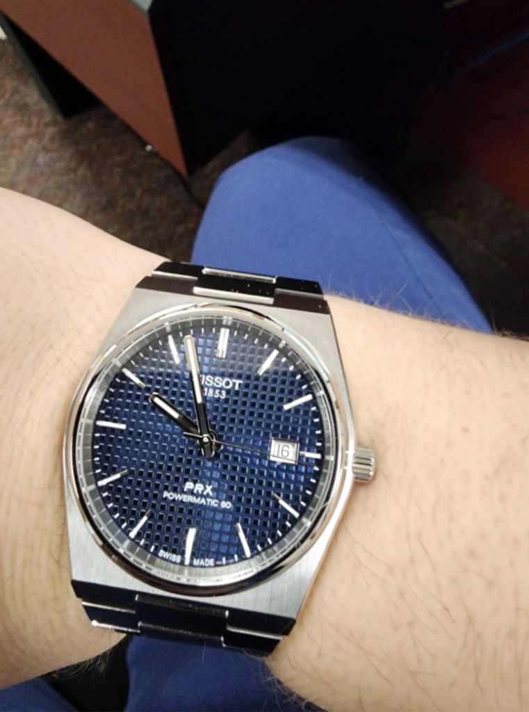 A wrist with a Tissot PRX Powermatic 80 watch featuring a blue textured dial and silver metal hands, displayed against a blurred indoor background. Date indicator shows the 16th. The band is metallic with a brushed finish.