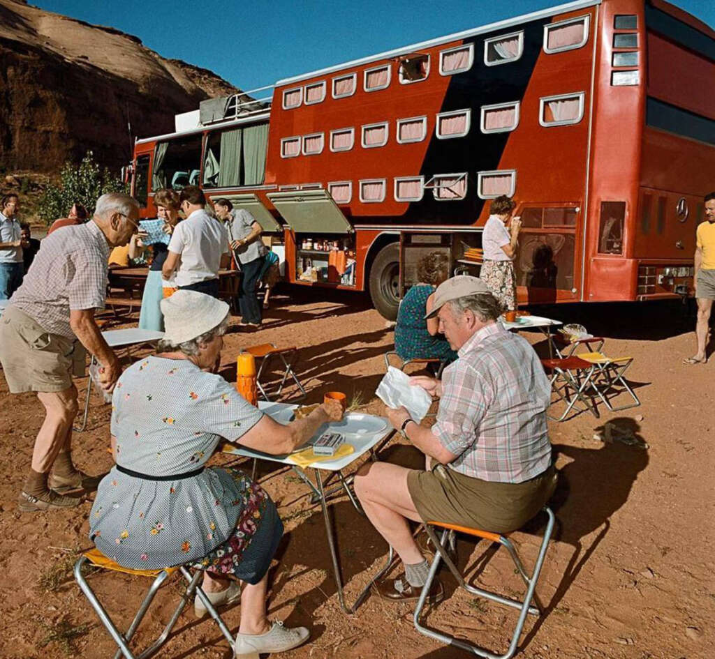 A large red double-decker bus parked in a desert area. Several people are sitting and standing around small tables, enjoying a meal. The group consists of mostly older adults, engaging in conversation under clear blue skies.