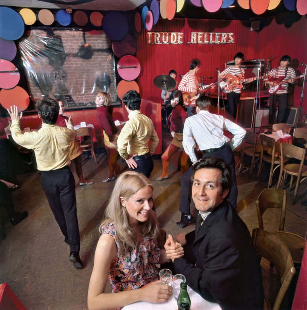 A lively 1960s nightclub scene with a couple seated at a table in the foreground. In the background, people dance near a stage with musicians wearing striped shirts. The club is colorful and energetic, with "TRUDE HELLER'S" as a sign on the wall.